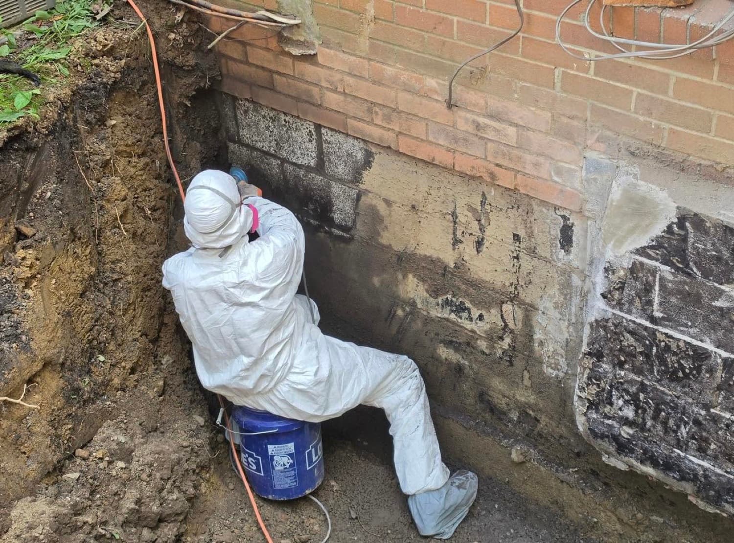 Aqua Solutions technician in a Tyvek suit and respirator hand-applying waterproofing sealer to an excavated brick-and-block foundation wall